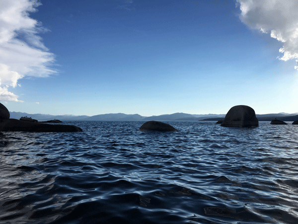 view of lake tahoe taken from kayak, punctuated with a few dark large round boulders. blue sky with a few white whispy clouds.