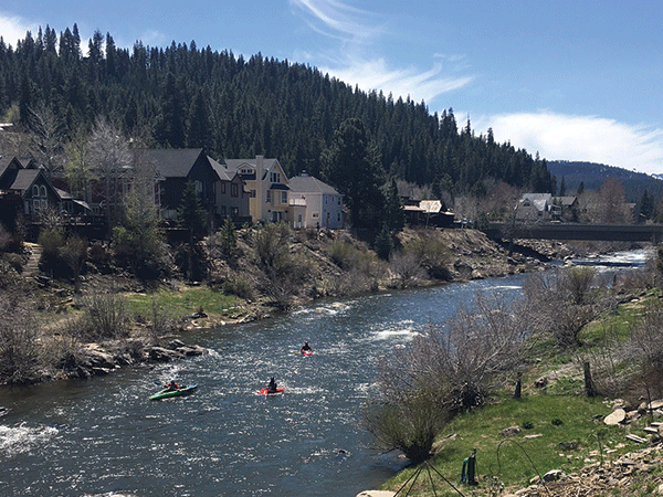 photo of 3 kayakers on the truckee river passing through downtown truckee with houses on left bank of river, bridge crossing river on right, dark green tree covered hill in background