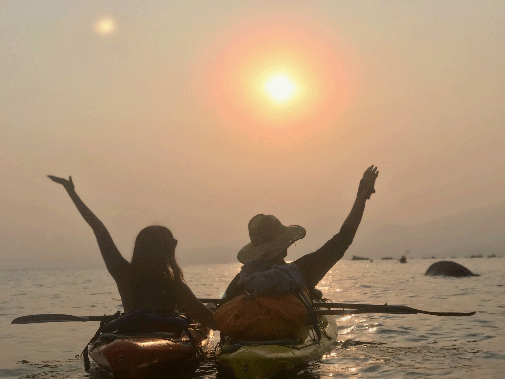 2 kayaks in lake tahoe, seated in them are a man and a woman, holding hands with smoke and sunset in background