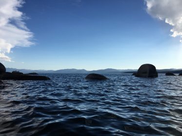 lake tahoe with deep blue water, a few dark round rocks, sky framed with 2 white fluffy clouds