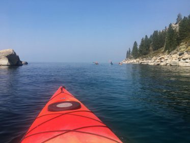 nose of red kayak looking out at 3 other kayaks in distance with rounded rocks of of lake tahoe's east shore and coastline