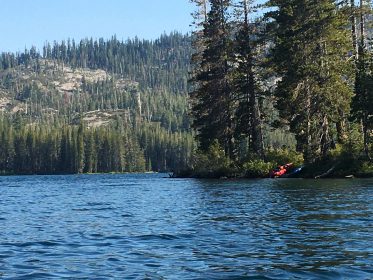 gold lake with open water on bottom half of photo, shoreline in center, 2 kayaks on right, and pine forest with granite hillside in background