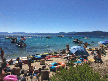 view of hidden beach on lake tahoe on busy july 4th weekend, filled with boats, kayaks, partiers on beach, american flags