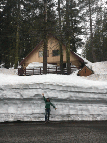 woman in green jacket in front of 10 foot snow wall blocking house in background in town of serene lakes california